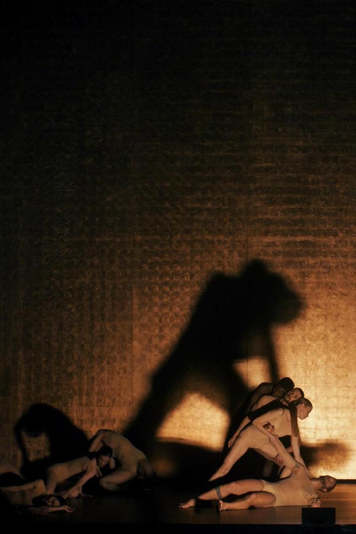 Mehrere Tänzer in beigen Kostümen auf einer Bühne mit goldenem Hintergrund.