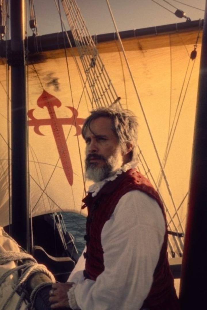 Ein Mann mit Bart und Weste steht auf einem Segelschiff mit dem Santiago-Kreuz.