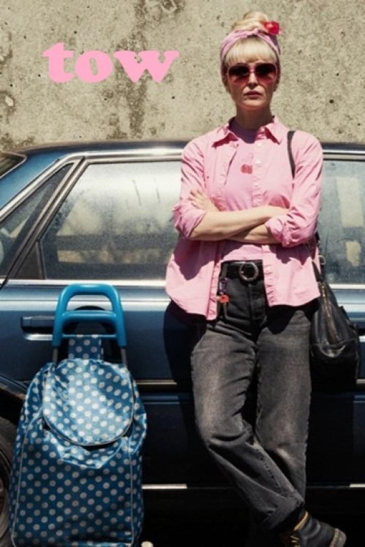 Eine Frau mit Sonnenbrille und rosa Hemd steht neben einem blauen Auto.