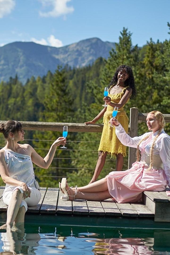 Drei Frauen stoßen mit blauen Getränken vor einer Bergkulisse an.