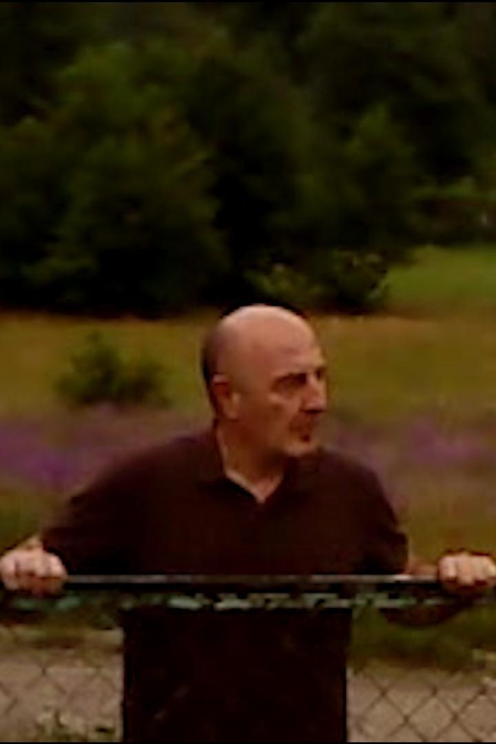 Ein Mann mit Glatze lehnt an einem Zaun und schaut nach rechts.