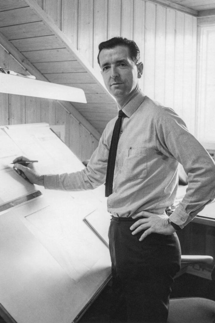 Ein Mann steht an einem Schreibtisch und zeichnet mit einem Stift auf ein Blatt Papier.