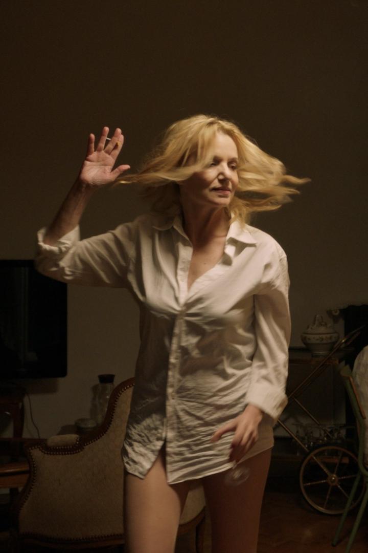 Eine Frau mit blonden Haaren trägt ein weißes Hemd und gestikuliert in einem dunklen Raum.