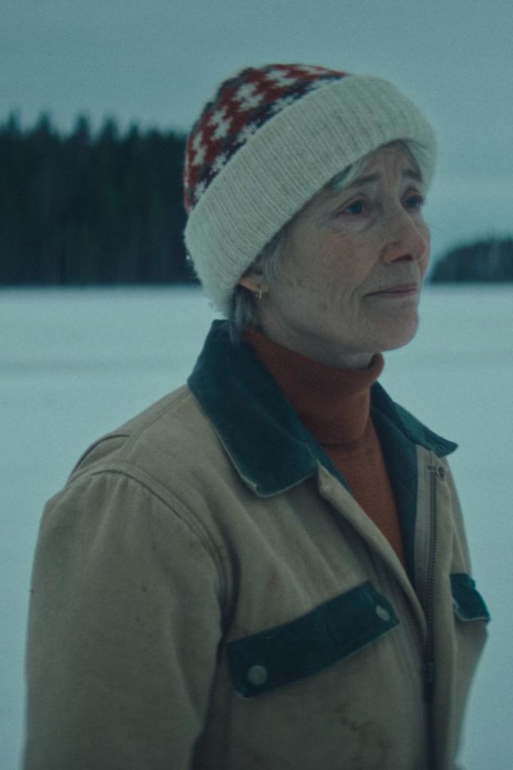 Eine ältere Frau mit Mütze und Jacke steht in einer verschneiten Landschaft.