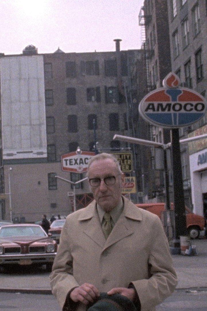 Ein älterer Mann mit Brille und Mantel steht vor einer Tankstelle in der Stadt.