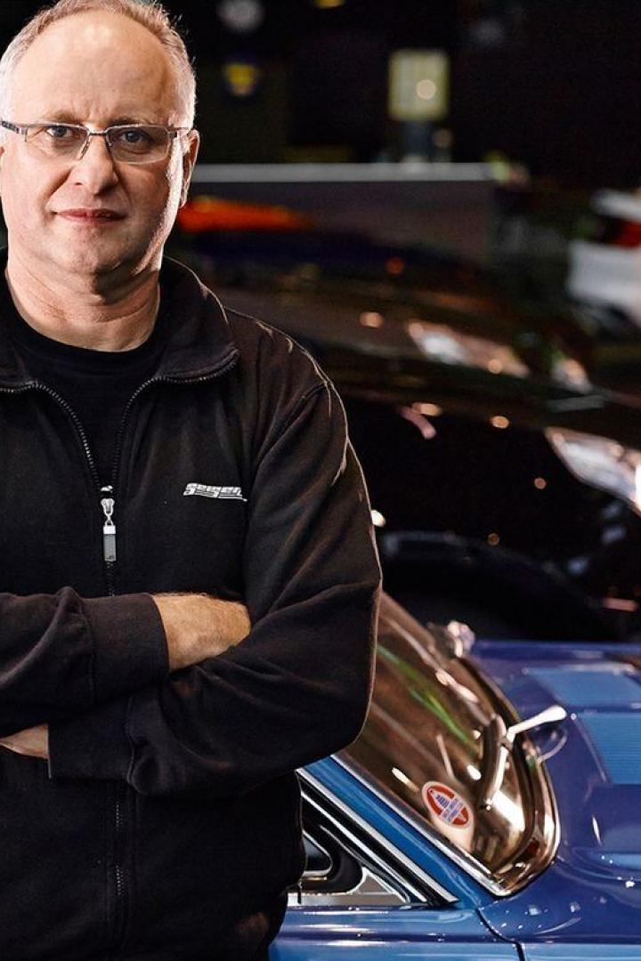 Ein Mann mit Brille steht vor einem blauen Oldtimer mit geöffneter Motorhaube.