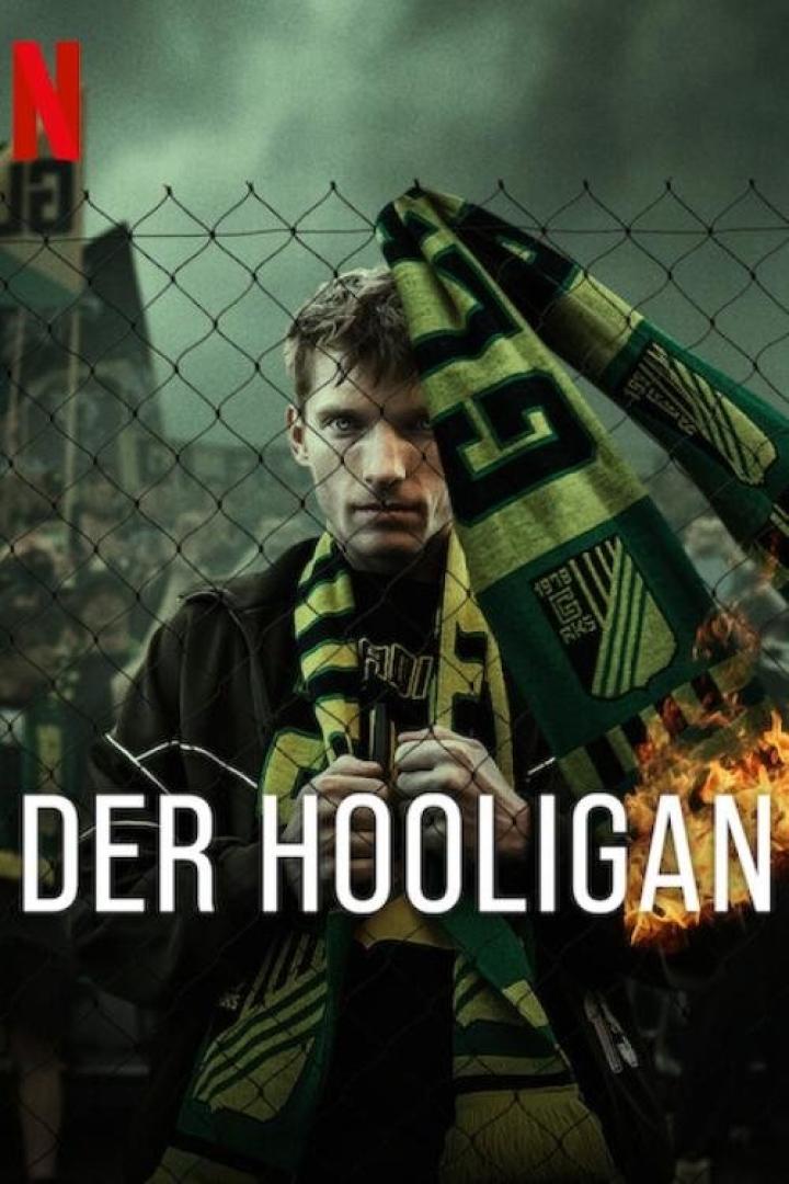 Ein Mann mit einem Schal steht vor einem Zaun, im Hintergrund das Logo von Netflix und der Titel „Der Hooligan“.
