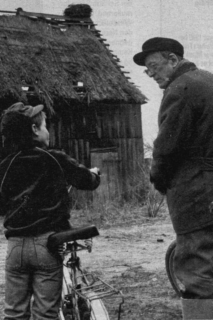 Ein Junge mit Fahrrad spricht mit einem älteren Mann vor einem Haus mit Strohdach.
