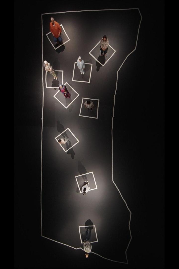 Eine Draufsicht zeigt Menschen, die in weißen Quadraten auf einer schwarzen Fläche stehen, die von einer weißen Linie umrandet ist.