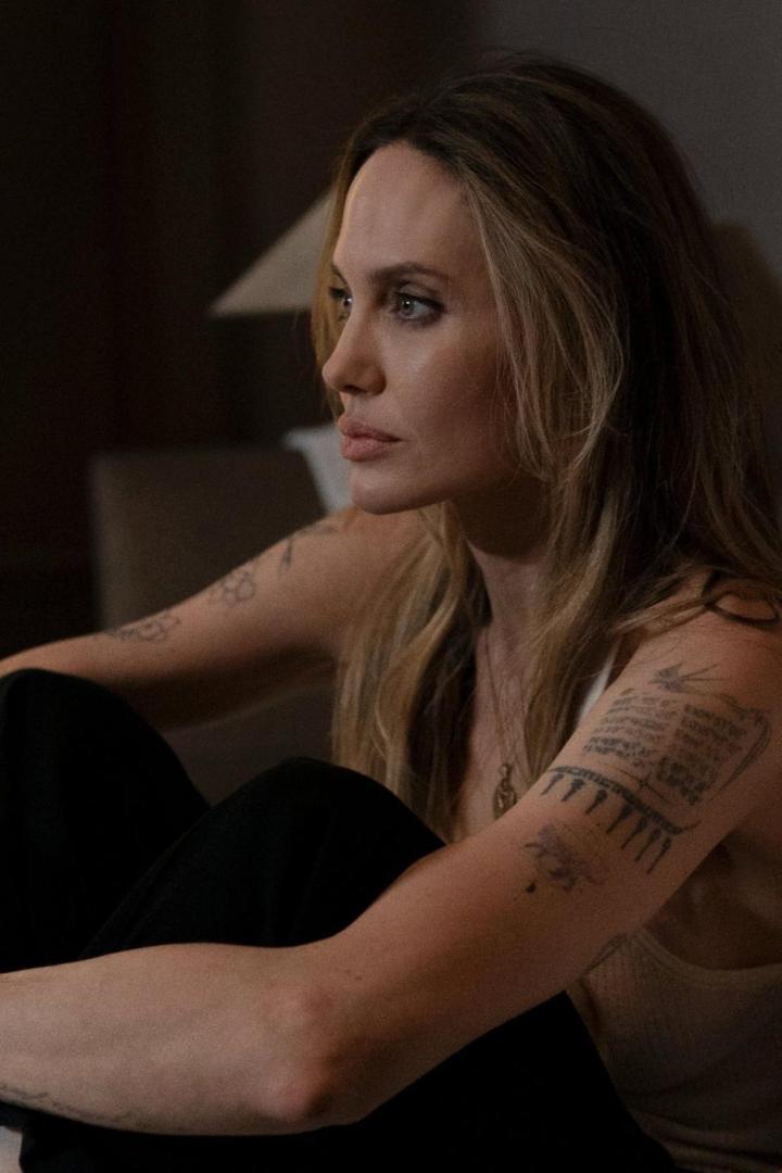 Angelina Jolie sitzt mit Tattoos auf den Armen und blickt zur Seite.