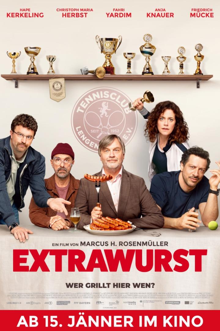 Hape Kerkeling, Christoph Maria Herbst und weitere Darsteller sitzen an einem Tisch mit Teller voller Grillwürsten unter Tennispokalen 