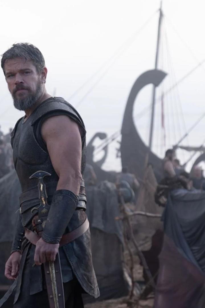 Matt Damon steht als Krieger Odysseus in Rüstung vor einem Lager mit antiken Schiffen.