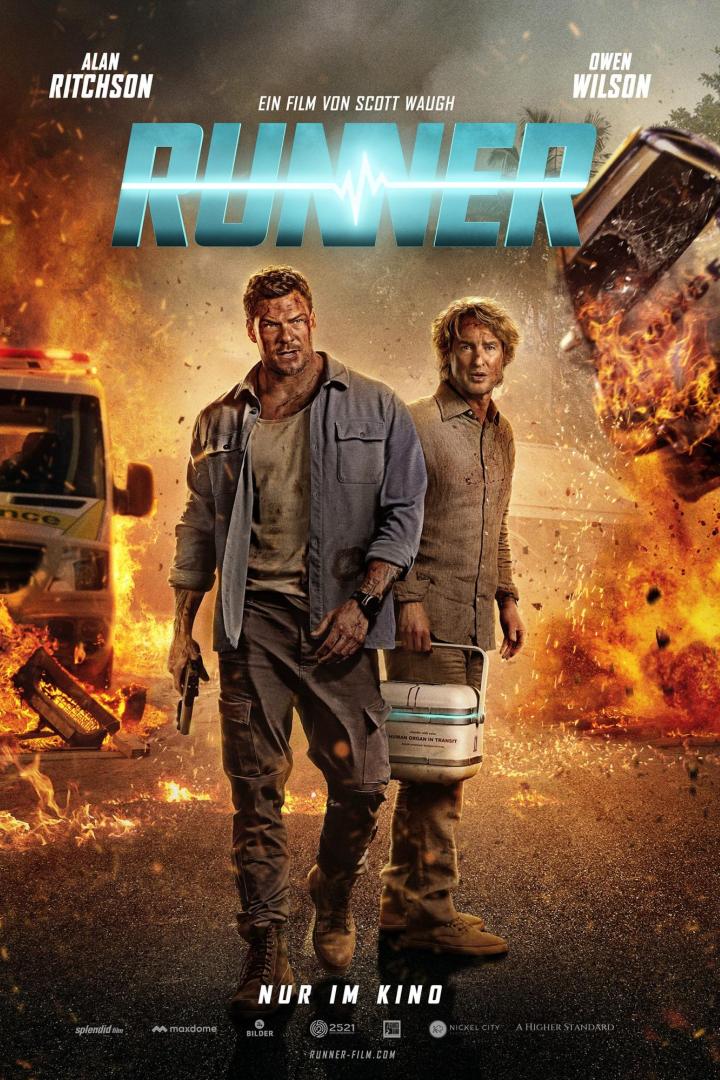 Alan Ritchson und Owen Wilson stehen vor brennenden und explodierenden  Autos, während sie einen Kühlbehälter für Organtransporte tragen.
