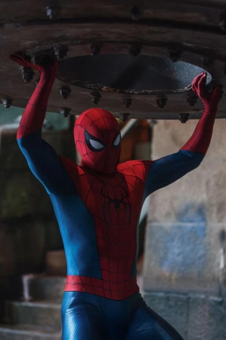 Spider-Man hebt in einem rot-blauen Kostüm eine schwere, metallische Konstruktion über seinen Kopf.