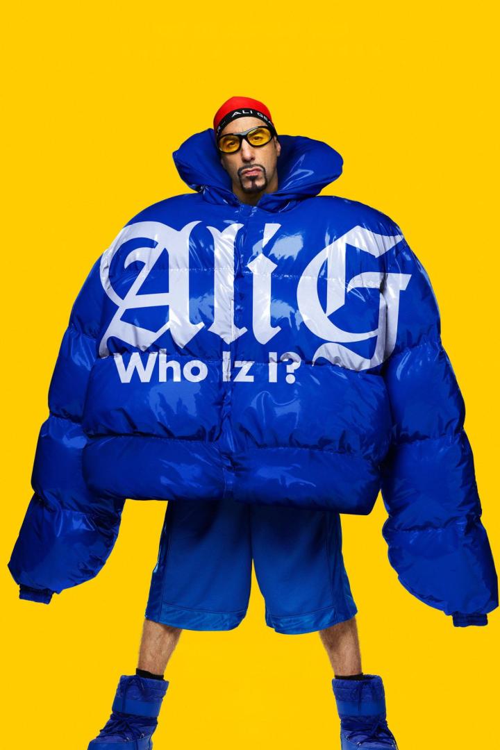 Ali G trägt eine überdimensionale blaue Daunenjacke mit der Aufschrift „Ali G Who Iz I?“ vor einem gelben Hintergrund.