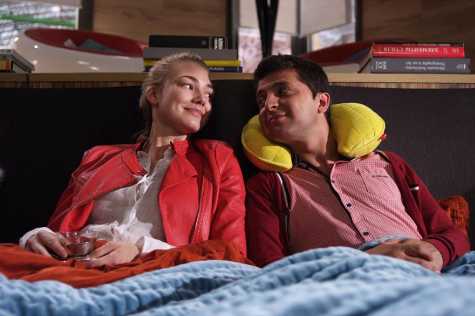 Ein Paar entspannt sich im Bett, die Frau hält ein Glas in der Hand, der Mann hat ein Nackenkissen.