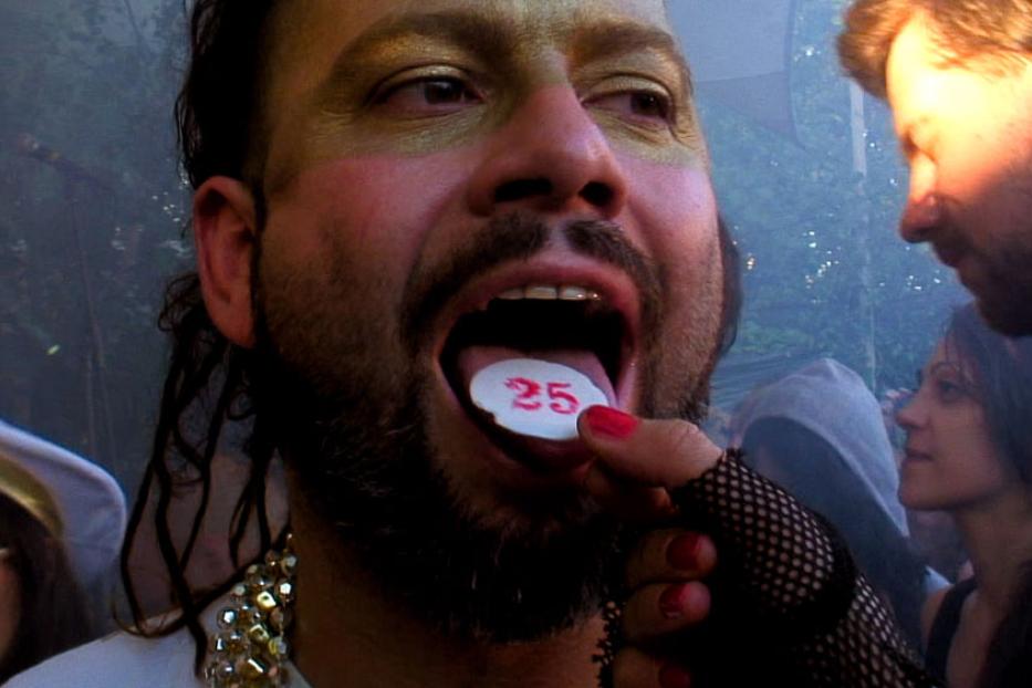 Ein Mann mit Make-up hält eine Pille mit der Zahl 25 auf seiner Zunge.