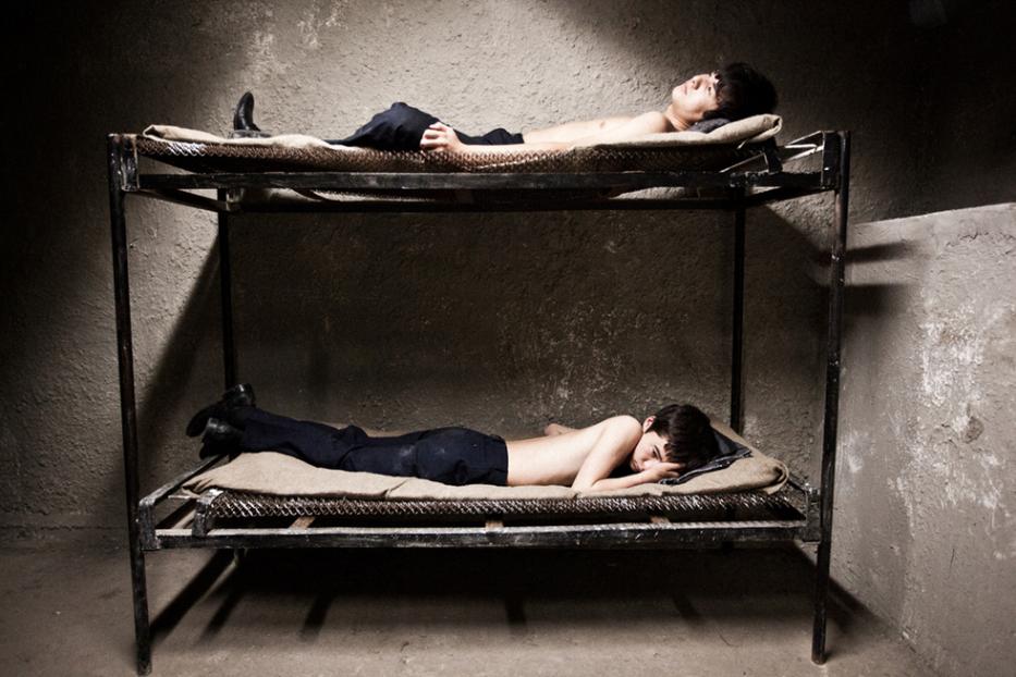 Zwei Männer liegen in einer kargen Zelle auf einer Doppelstockliege.