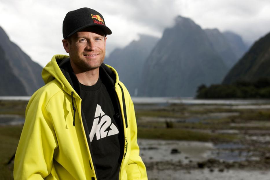 Ein Mann mit Kappe und gelber Jacke vor einer Berglandschaft.