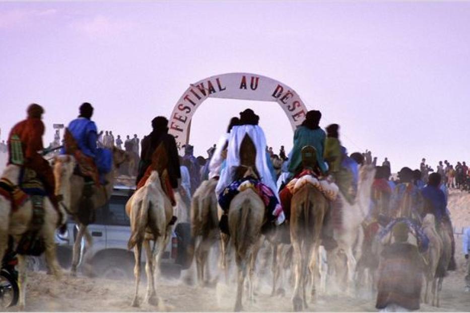 Eine Gruppe von Menschen reitet auf Kamelen durch einen Bogen mit der Aufschrift „Festival au Desert“.