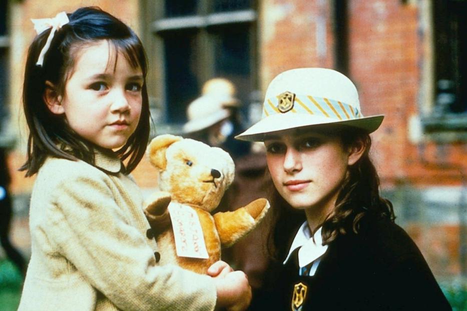 Zwei junge Mädchen, eines mit einem Teddybär, stehen vor einem Backsteingebäude.