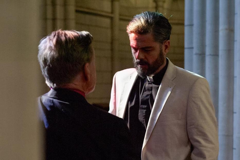 Ein bärtiger Mann im Priesterhemd und hellem Sakko unterhält sich mit einer anderen Person.