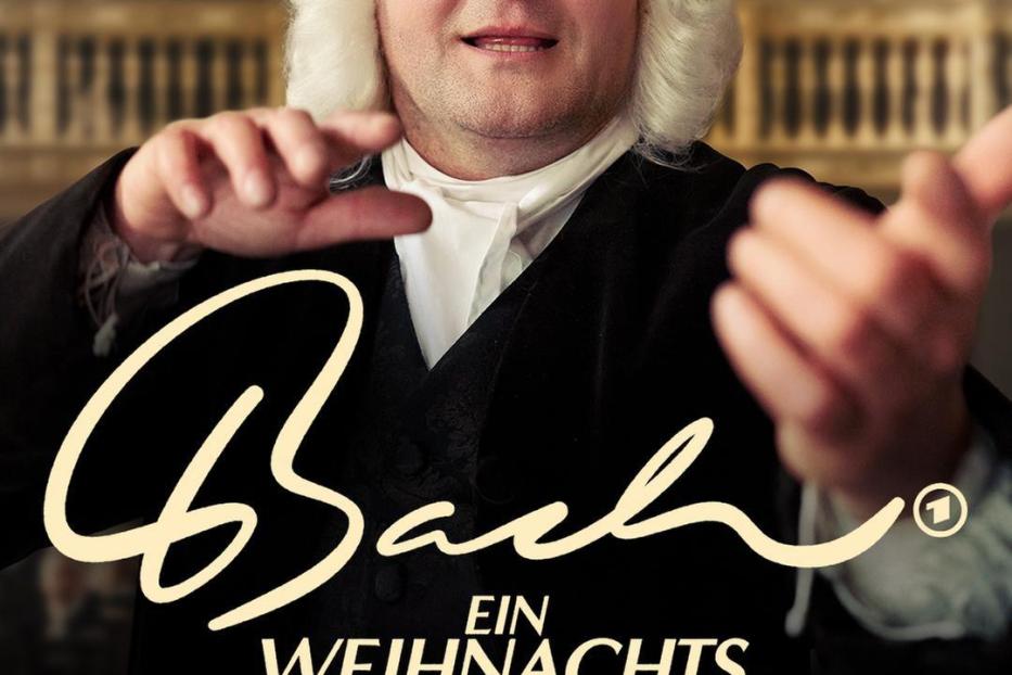 Ein Mann in historischer Kleidung und Perücke vor dem Hintergrund des Films „Bach – Ein Weihnachtswunder“.