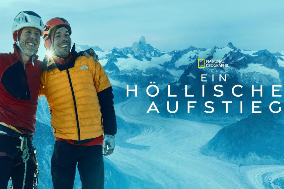 Zwei Bergsteiger umarmen sich vor einer verschneiten Berglandschaft.