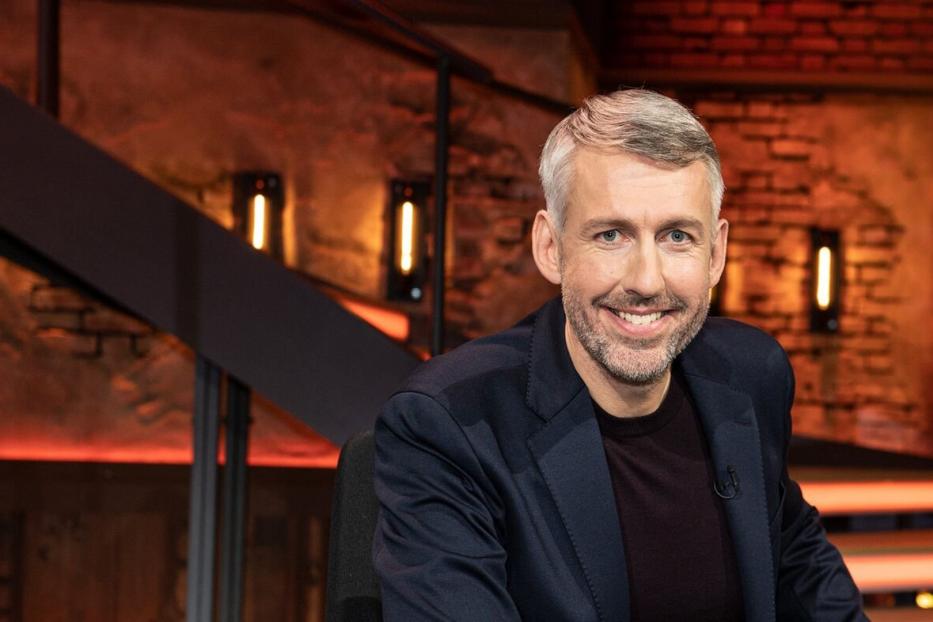 Ein Mann mit grauem Haar und Bart lächelt in die Kamera, im Hintergrund eine Treppe.