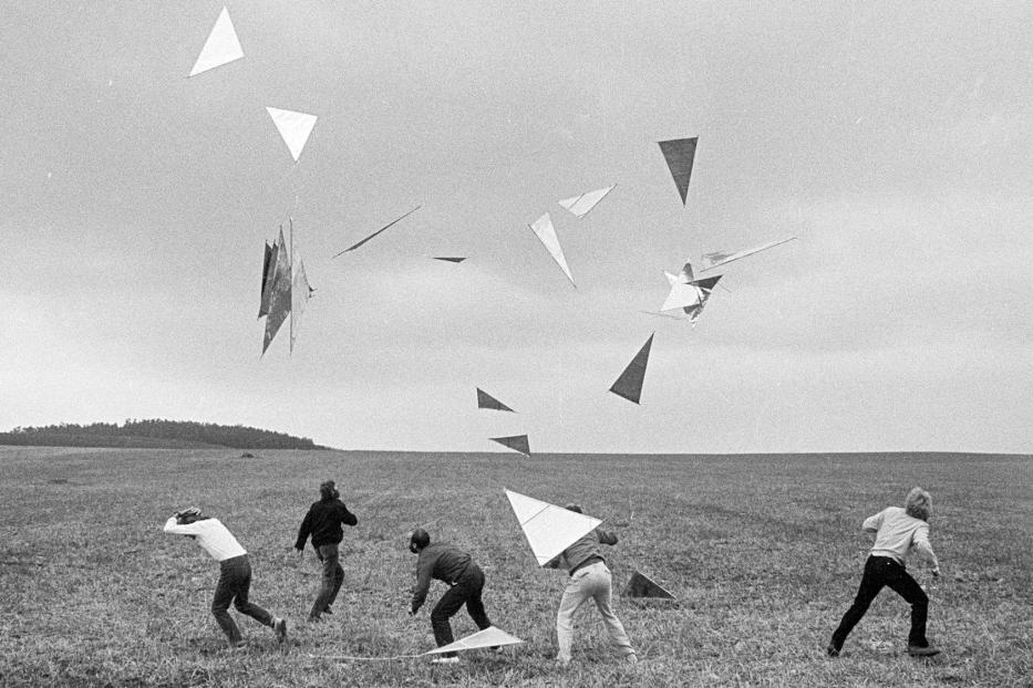 Fünf Personen rennen auf einem Feld unter einem Schwarm dreieckiger Drachen umher.