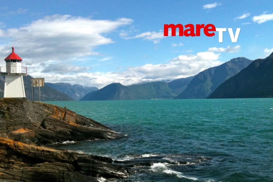 Ein Leuchtturm steht an der Küste eines Fjords mit Bergen im Hintergrund.