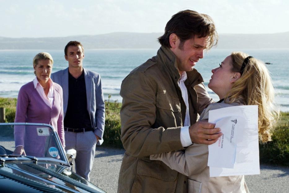 Ein Paar umarmt sich innig vor einer Küstenlandschaft, während zwei weitere Personen im Hintergrund stehen.