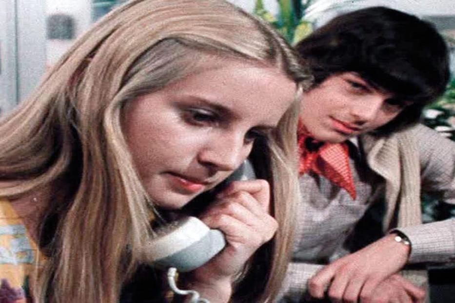 Eine junge Frau telefoniert, während ein junger Mann ihr über die Schulter schaut.