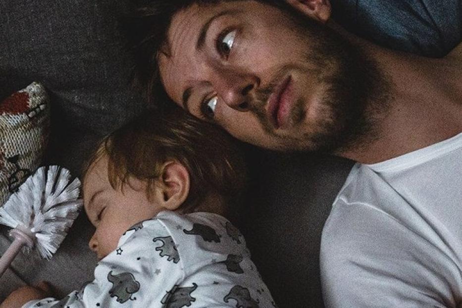 Ein Vater liegt neben seinem schlafenden Kind auf einem Sofa.