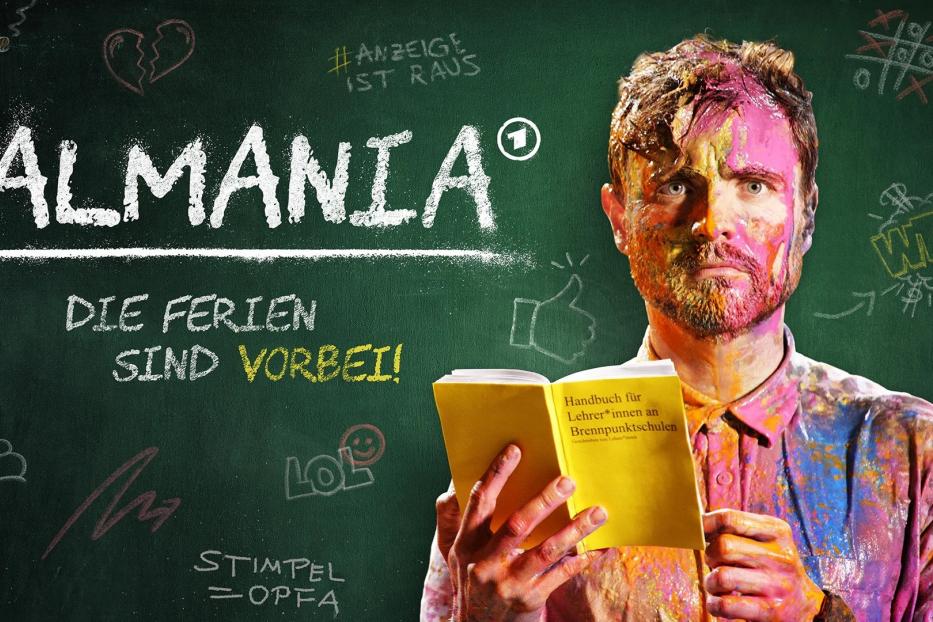 Ein mit Farbe beschmierter Mann hält ein Buch mit dem Titel „Handbuch für Lehrer*innen an Brennpunktschulen“ vor einer Tafel mit der Aufschrift „Almania“.