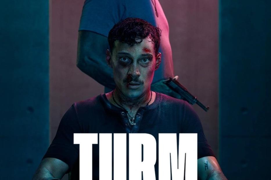 Ein Mann mit Handschellen sitzt auf einem Stuhl, während ein älterer Mann mit einer Waffe hinter ihm steht. Titel: „Turm Schatten“.