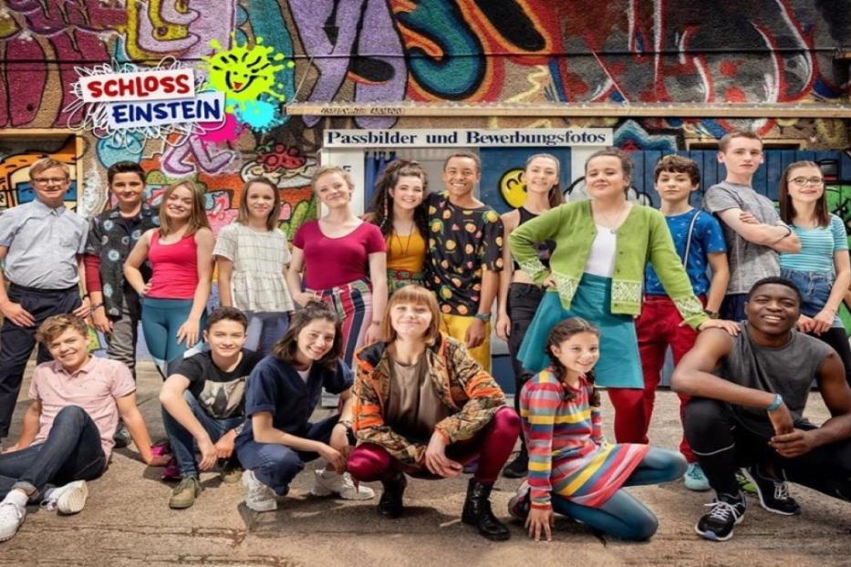 Eine Gruppe Jugendlicher steht vor einer mit Graffiti bemalten Wand mit dem „Schloss Einstein“-Logo.
