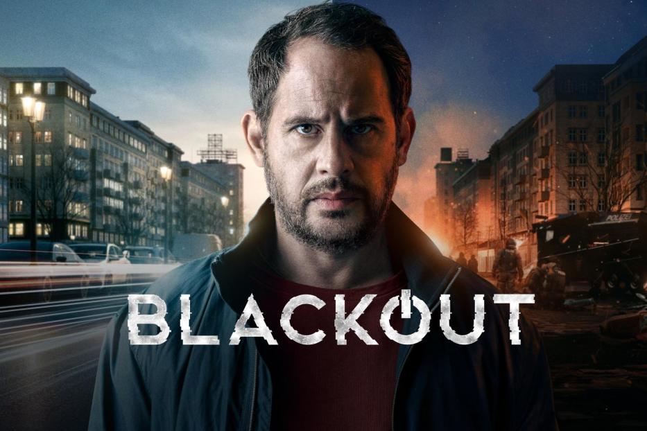 Ein Mann vor dem Hintergrund des Posters für die Fernsehserie „Blackout“.