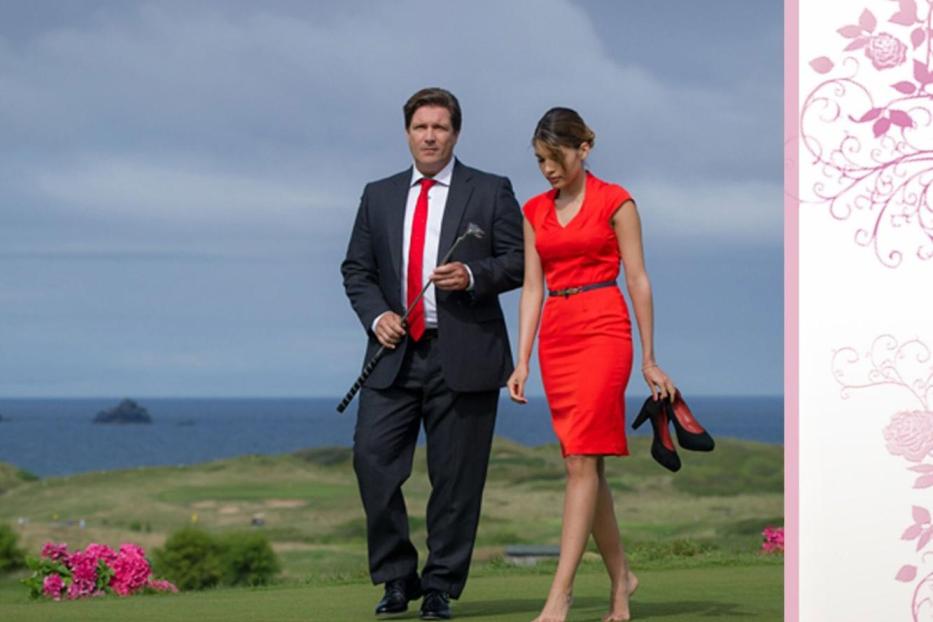 Ein Mann im Anzug und eine Frau im roten Kleid gehen barfuß auf einem Golfplatz am Meer entlang.