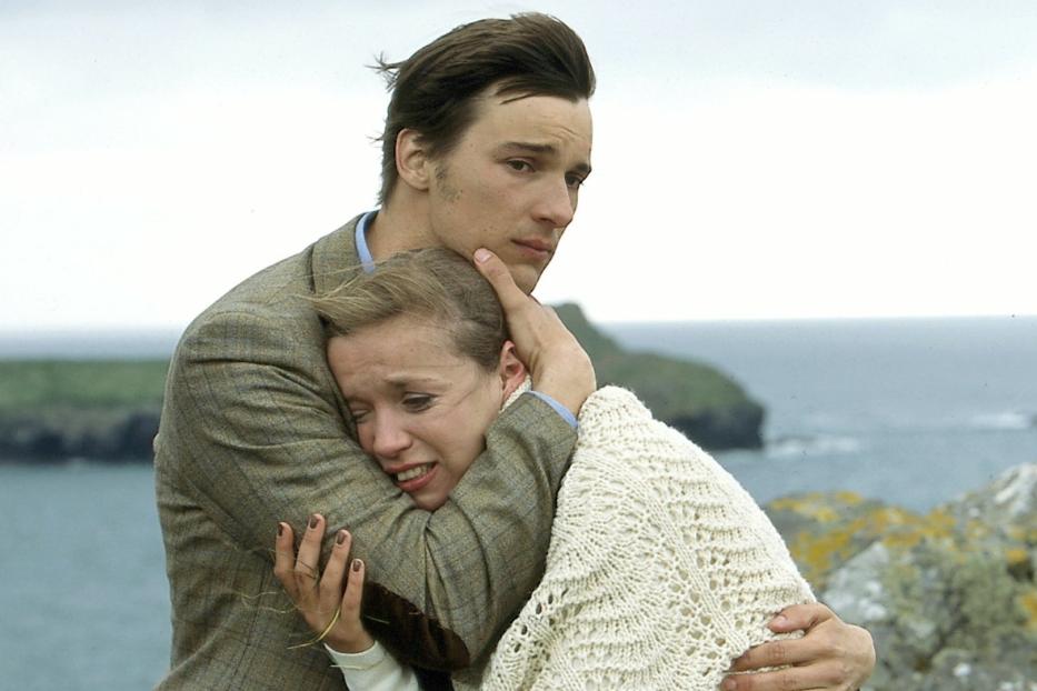 Ein Mann umarmt eine weinende Frau an einer Küste.