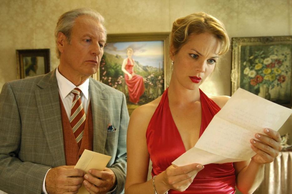 Eine Frau im roten Kleid liest einen Brief, während ein Mann im Anzug daneben steht.