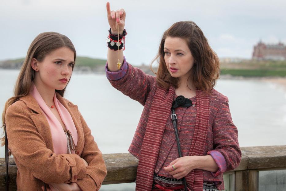 Zwei Frauen stehen an einer Küste, eine hält ein Pendel in der Hand.