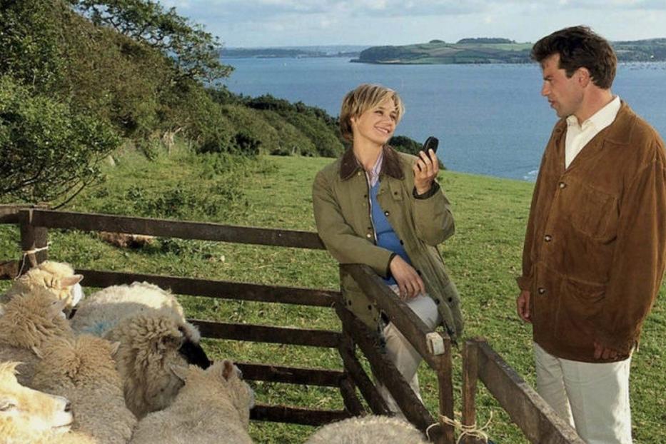 Eine Frau telefoniert auf einer Weide mit Schafen, während ein Mann daneben steht.