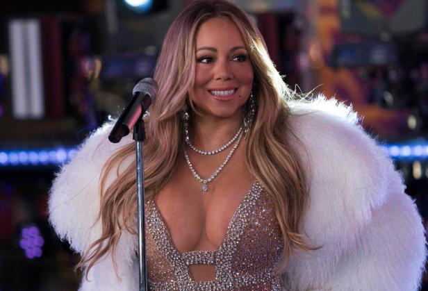 Mariah Carey steht lächelnd mit Glitzerkleid und weißem Fellmantel vor einem Mikrofonständer