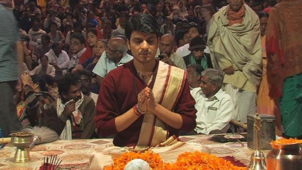 Ein Mann sitzt in traditioneller Kleidung mit gefalteten Händen vor einer Menschenmenge.