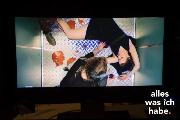 Zwei Frauen liegen auf einem Boden mit zerbrochenen Puppenköpfen und Blutattrappen.