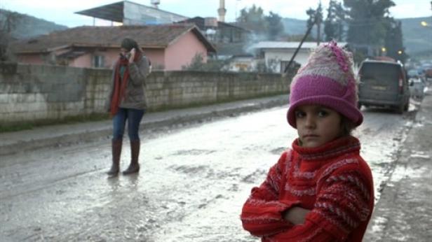 Ein ernstes Mädchen mit Mütze steht auf einer schlammigen Straße, während eine Frau telefoniert.