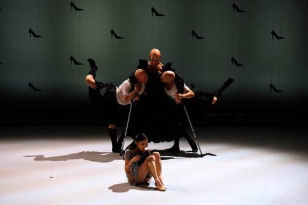 Eine Frau sitzt am Boden, während Tänzer eine Performance mit High Heels im Hintergrund aufführen.