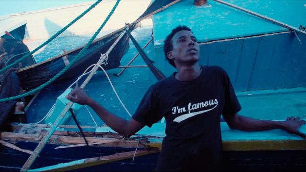 Ein junger Mann steht vor einem beschädigten, blau gestrichenen Boot.