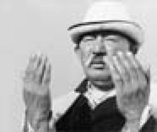 Ein Mann mit Hut und Schnurrbart hält die Hände vor das Gesicht.